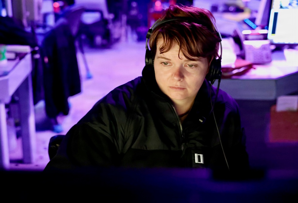 A person wearing a black jacket and over-ear headphones is focused on a computer screen in a dimly lit workspace. The scene is illuminated by a blue-purple ambient light, casting a cool tone across the environment. The individual appears to be engaged in concentrated work at what looks like a multi-monitor workstation.