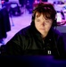 A person wearing a black jacket and over-ear headphones is focused on a computer screen in a dimly lit workspace. The scene is illuminated by a blue-purple ambient light, casting a cool tone across the environment. The individual appears to be engaged in concentrated work at what looks like a multi-monitor workstation.