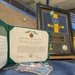The image shows a military awards display for Captain Scott L. Leier, featuring a Department of the Army Commendation Medal certificate, a framed Sixty-Eighth Troop Command guidon/flag plaque, and a blue uniform item with an embroidered insignia. The items are arranged on a table, likely at a formal military ceremony or recognition event. The certificate is signed by Colonel Cody J. Volk, Commanding, and is dated September 12, 2026, covering service from 01 February 2024 to 30 September 2026.