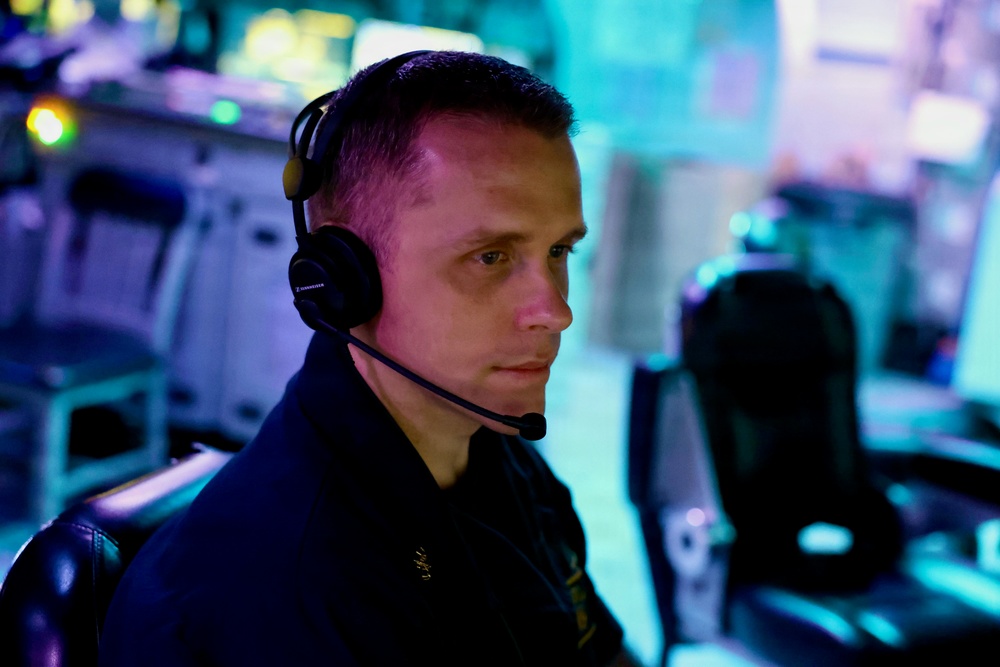 A focused individual wearing a Sennheiser communications headset is seated at what appears to be a command or operations center workstation. The environment is illuminated with blue and teal ambient lighting, suggesting a tactical, military, or broadcast control room setting. The subject is wearing a dark uniform with a visible insignia and appears to be actively monitoring or communicating.