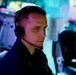 A focused individual wearing a Sennheiser communications headset is seated at what appears to be a command or operations center workstation. The environment is illuminated with blue and teal ambient lighting, suggesting a tactical, military, or broadcast control room setting. The subject is wearing a dark uniform with a visible insignia and appears to be actively monitoring or communicating.