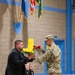 A U.S. Army soldier in OCP (Operational Camouflage Pattern) uniform is presenting a bouquet of red and yellow roses to a civilian man in a black suit during what appears to be a military ceremony held in an armory or gymnasium. Two young boys dressed in formal black attire are seated behind the civilian, watching the exchange. Flags including the U.S. flag and what appears to be a state flag are displayed on the wall in the background.