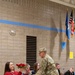 A military service member in camouflage uniform is presenting red roses to a woman seated in a folding chair during what appears to be a formal military ceremony held in a gymnasium or community center. Several other individuals, including children and adults, are seated in a row of chairs nearby, and multiple flags including the American flag are displayed on the wall in the background. The scene suggests a homecoming, recognition, or farewell ceremony.