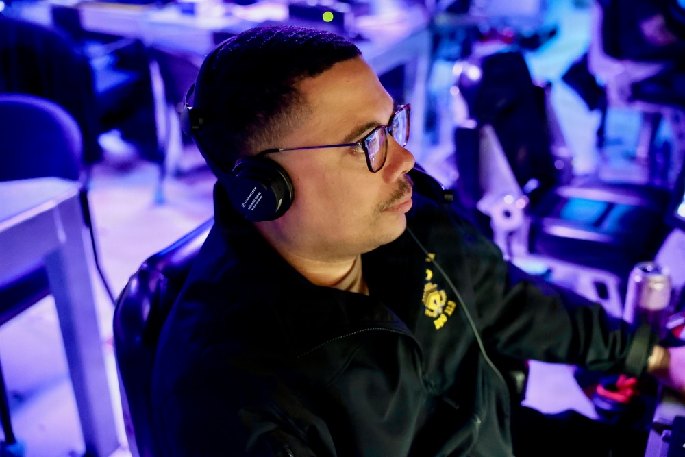 A man wearing black over-ear headphones and glasses is seated at what appears to be an esports or gaming event, focused intently on something off-camera. He is wearing a black jacket with a yellow logo/emblem on the chest. The environment is lit with blue ambient lighting typical of competitive gaming venues.