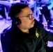 A man wearing black over-ear headphones and glasses is seated at what appears to be an esports or gaming event, focused intently on something off-camera. He is wearing a black jacket with a yellow logo/emblem on the chest. The environment is lit with blue ambient lighting typical of competitive gaming venues.