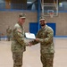 Two U.S. Army soldiers in OCP (Operational Camouflage Pattern) uniforms are shaking hands while jointly holding a certificate or award document in what appears to be a military gymnasium. The ceremony appears to be a formal award or commendation presentation. A third soldier stands at attention in the background.