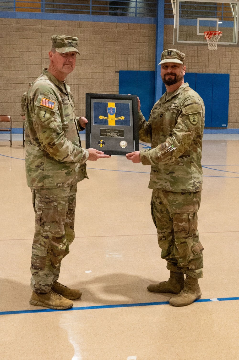 Two U.S. Army soldiers in MultiCam/OCP camouflage uniforms are posing together in a gymnasium while jointly holding a framed military plaque or shadow box. The framed item appears to contain a unit crest with blue and yellow colors, along with coins and an engraved plate. This appears to be a formal military recognition or farewell ceremony.
