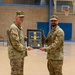 Two U.S. Army soldiers in MultiCam/OCP camouflage uniforms are posing together in a gymnasium while jointly holding a framed military plaque or shadow box. The framed item appears to contain a unit crest with blue and yellow colors, along with coins and an engraved plate. This appears to be a formal military recognition or farewell ceremony.