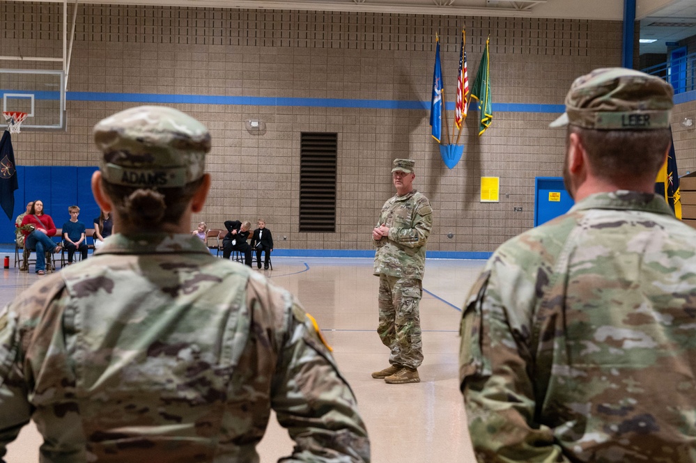 A military ceremony is taking place inside a gymnasium, with a senior soldier addressing formation members from the front. Two soldiers in the foreground, identified by name tapes 'ADAMS' and 'LEER', are facing a speaker standing at attention in the center of the gym. Flags including the American flag and what appear to be state and unit flags are displayed in the background.