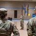 A military ceremony is taking place inside a gymnasium, with a senior soldier addressing formation members from the front. Two soldiers in the foreground, identified by name tapes 'ADAMS' and 'LEER', are facing a speaker standing at attention in the center of the gym. Flags including the American flag and what appear to be state and unit flags are displayed in the background.