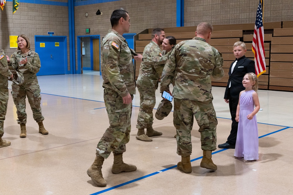 A military ceremony is taking place inside what appears to be a school gymnasium. Several soldiers in Army Combat Uniforms (ACU/OCP pattern) are gathered, with one soldier appearing to receive a pin or badge from another. Two children dressed formally — a boy in a tuxedo and a girl in a lavender dress — are present, suggesting a promotion or award ceremony involving family members.