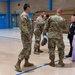 A military ceremony is taking place inside what appears to be a school gymnasium. Several soldiers in Army Combat Uniforms (ACU/OCP pattern) are gathered, with one soldier appearing to receive a pin or badge from another. Two children dressed formally — a boy in a tuxedo and a girl in a lavender dress — are present, suggesting a promotion or award ceremony involving family members.