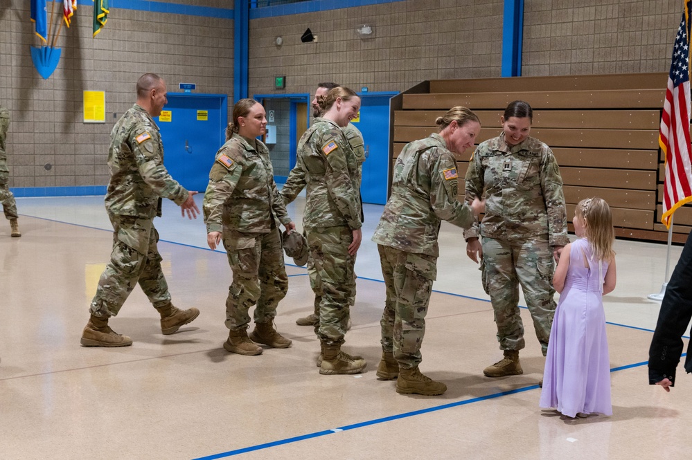 A military ceremony taking place inside a gymnasium, likely a deployment or homecoming event. Several U.S. Army soldiers in MultiCam/OCP uniforms are gathered, with one female soldier interacting warmly with a young girl in a lavender dress. An American flag and what appears to be a unit guidon are visible in the background.