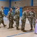 A military ceremony taking place inside a gymnasium, likely a deployment or homecoming event. Several U.S. Army soldiers in MultiCam/OCP uniforms are gathered, with one female soldier interacting warmly with a young girl in a lavender dress. An American flag and what appears to be a unit guidon are visible in the background.