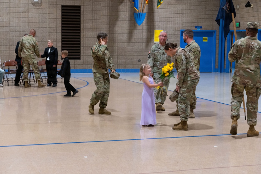 A military ceremony or event is taking place inside a gymnasium or indoor facility. A young girl in a lavender dress is presenting yellow flowers to a female soldier who is bending down to interact with her. Multiple soldiers in camouflage uniforms are present, along with civilians including children in formal attire.