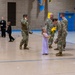 A military ceremony or event is taking place inside a gymnasium or indoor facility. A young girl in a lavender dress is presenting yellow flowers to a female soldier who is bending down to interact with her. Multiple soldiers in camouflage uniforms are present, along with civilians including children in formal attire.