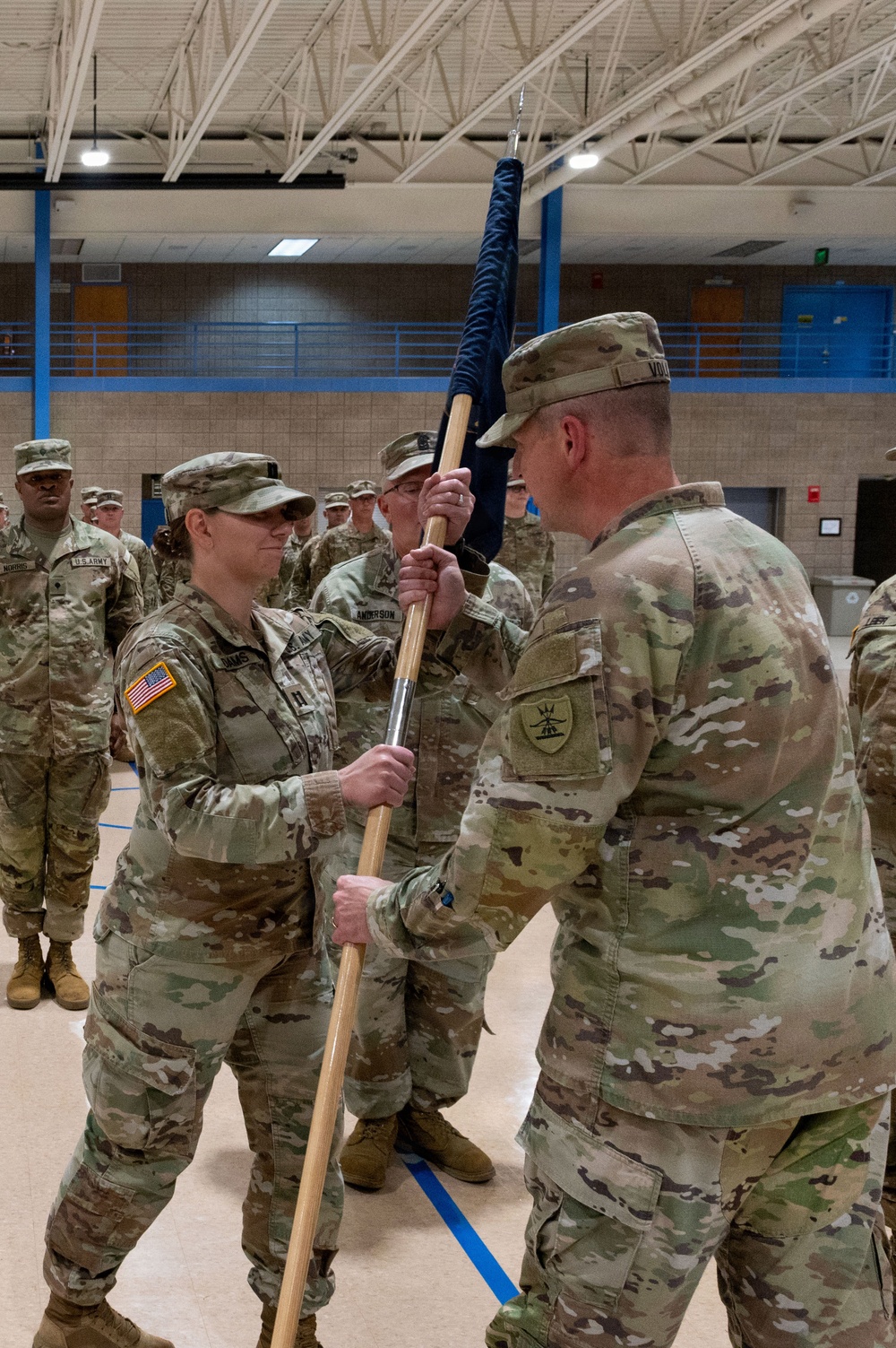A U.S. Army change of command ceremony is taking place inside a military gymnasium or armory. A female soldier is receiving or passing a unit guidon (furled flag on a staff) to/from another soldier, which is the central ritual of a change of command ceremony. Multiple soldiers in OCP (Operational Camouflage Pattern) uniforms are standing in formation in the background.
