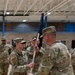 A U.S. Army change of command ceremony is taking place inside a military gymnasium or armory. A female soldier is receiving or passing a unit guidon (furled flag on a staff) to/from another soldier, which is the central ritual of a change of command ceremony. Multiple soldiers in OCP (Operational Camouflage Pattern) uniforms are standing in formation in the background.