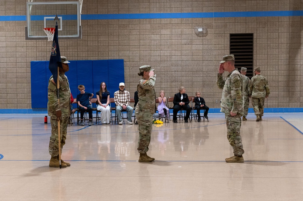 A military ceremony is taking place inside a gymnasium, likely a change of command or promotion ceremony. Two soldiers in the foreground are exchanging salutes while a third soldier holds a unit guidon flag. Civilian family members, including children dressed formally, are seated in the background as observers.
