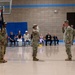 A military ceremony is taking place inside a gymnasium, likely a change of command or promotion ceremony. Two soldiers in the foreground are exchanging salutes while a third soldier holds a unit guidon flag. Civilian family members, including children dressed formally, are seated in the background as observers.