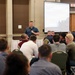 A speaker addresses a seated audience in a conference or banquet hall setting. The presenter stands at the front holding a microphone, with a projected presentation screen visible behind him displaying a slide titled 'From the Desk of the SP'. The audience consists of approximately 20-30 individuals seated at round and rectangular tables.