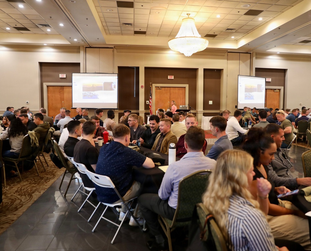 A large conference or seminar is taking place in a hotel ballroom with approximately 80-100 attendees seated at round and rectangular tables. A female presenter stands at the front of the room near a podium, with two large projection screens displaying what appears to be a slideshow presentation. The room features a prominent chandelier, recessed lighting, and an American flag near the front.
