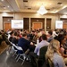 A large conference or seminar is taking place in a hotel ballroom with approximately 80-100 attendees seated at round and rectangular tables. A female presenter stands at the front of the room near a podium, with two large projection screens displaying what appears to be a slideshow presentation. The room features a prominent chandelier, recessed lighting, and an American flag near the front.