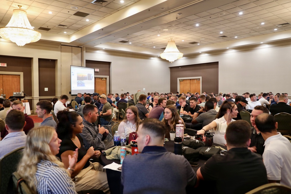 A large group of people, predominantly young adults, are seated at round tables in a hotel ballroom or conference center. A presentation is being displayed on a projection screen at the front of the room. The event appears to be a seminar, conference, or training session with approximately 100 or more attendees.