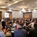 A large group of people, predominantly young adults, are seated at round tables in a hotel ballroom or conference center. A presentation is being displayed on a projection screen at the front of the room. The event appears to be a seminar, conference, or training session with approximately 100 or more attendees.