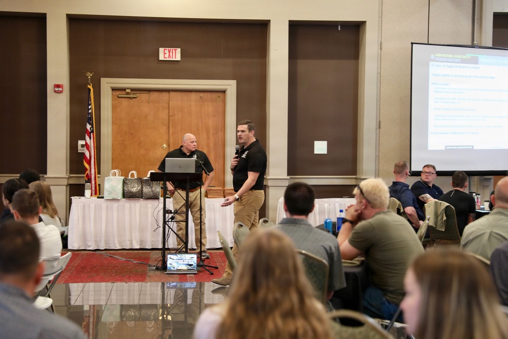 Two men in matching black polo shirts and khaki pants are presenting at what appears to be a formal conference or seminar in a banquet hall. One presenter holds a microphone while the other stands near a laptop at a podium. An audience of approximately 20+ attendees is seated at round tables facing the presenters, with a projection screen displaying a slide visible on the right.
