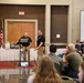 Two men in matching black polo shirts and khaki pants are presenting at what appears to be a formal conference or seminar in a banquet hall. One presenter holds a microphone while the other stands near a laptop at a podium. An audience of approximately 20+ attendees is seated at round tables facing the presenters, with a projection screen displaying a slide visible on the right.