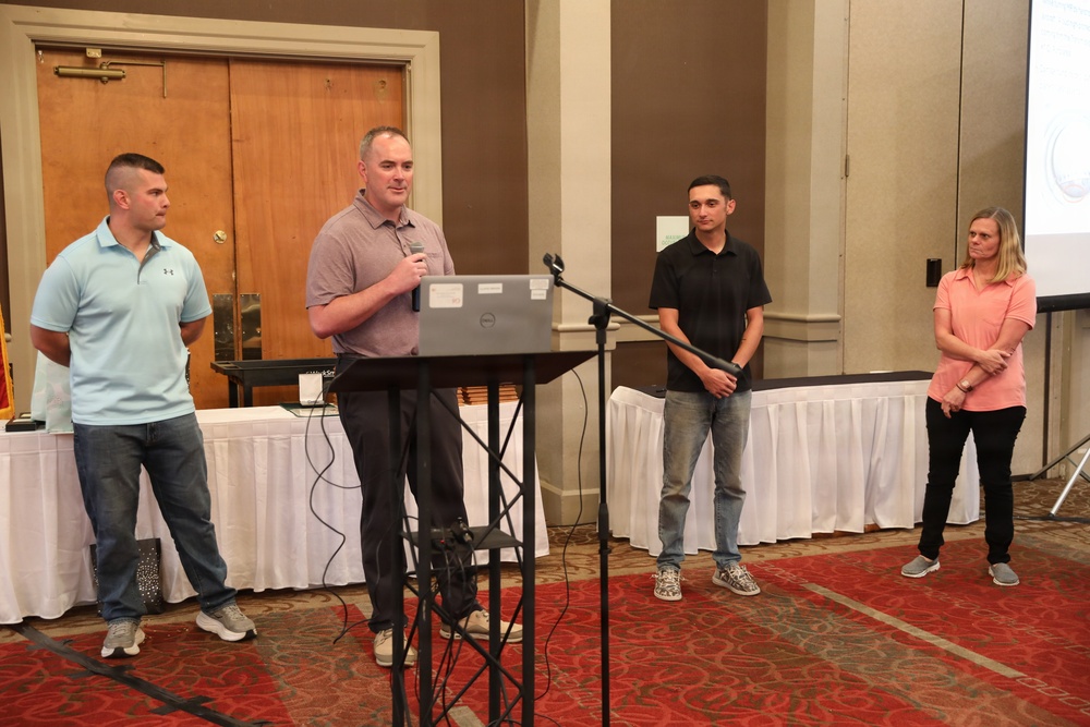 Four individuals are standing at the front of a banquet or conference room, with one person speaking at a podium equipped with a laptop and microphone. The setting appears to be a formal presentation or award ceremony in a hotel or event venue. A projection screen is visible in the background displaying content.
