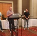 Four individuals are standing at the front of a banquet or conference room, with one person speaking at a podium equipped with a laptop and microphone. The setting appears to be a formal presentation or award ceremony in a hotel or event venue. A projection screen is visible in the background displaying content.