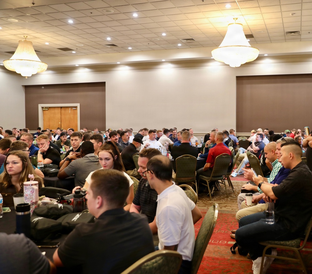 A large gathering of people seated at round tables in a hotel or convention center ballroom. The room is filled with dozens of attendees, suggesting a conference, seminar, or organized event. The space features classic chandelier lighting and a drop ceiling typical of banquet hall venues.