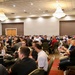 A large gathering of people seated at round tables in a hotel or convention center ballroom. The room is filled with dozens of attendees, suggesting a conference, seminar, or organized event. The space features classic chandelier lighting and a drop ceiling typical of banquet hall venues.