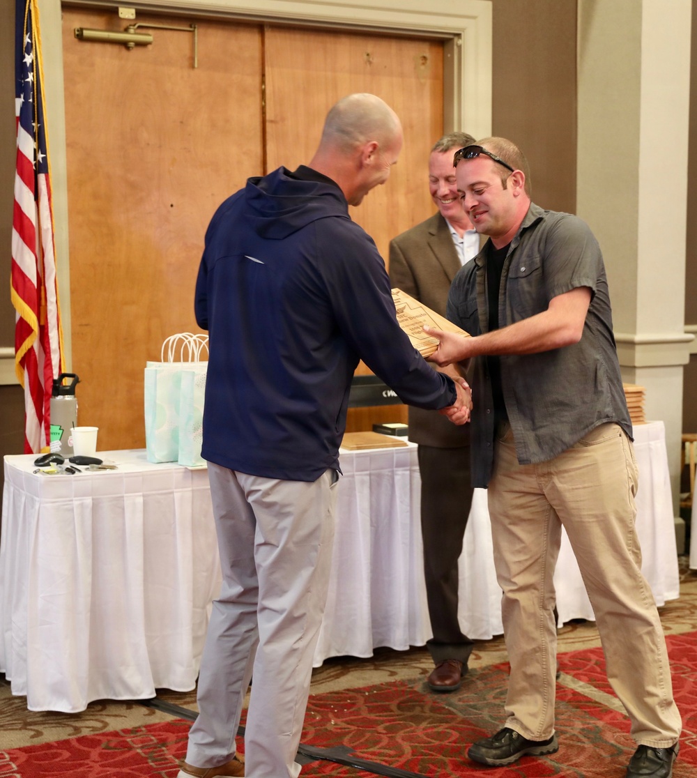 Two men are shaking hands while exchanging what appears to be a wooden plaque or award at a formal ceremony. A third man in a suit stands in the background smiling. The setting appears to be a banquet hall or conference room with an American flag visible on the left.