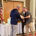 Two men are shaking hands while exchanging what appears to be a wooden plaque or award at a formal ceremony. A third man in a suit stands in the background smiling. The setting appears to be a banquet hall or conference room with an American flag visible on the left.