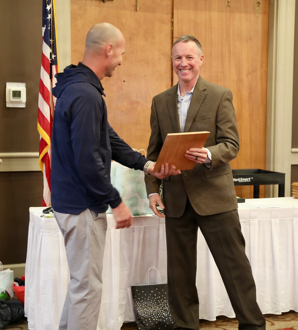 Two men are engaged in what appears to be an award or recognition ceremony, with one man in a brown blazer smiling broadly while holding a wooden plaque. They appear to be shaking hands or exchanging the plaque in a formal indoor setting. An American flag is visible in the background, suggesting an official or civic event.