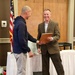 Two men are engaged in what appears to be an award or recognition ceremony, with one man in a brown blazer smiling broadly while holding a wooden plaque. They appear to be shaking hands or exchanging the plaque in a formal indoor setting. An American flag is visible in the background, suggesting an official or civic event.