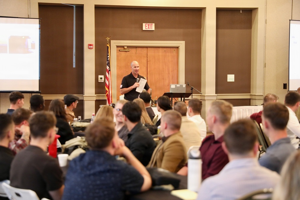 A speaker dressed in a black polo shirt stands at the front of a conference or seminar room, holding papers and addressing a seated audience. The room contains projection screens on both sides displaying presentation content, and an American flag is visible near the rear door. The audience appears to be a mixed group of young adults seated in rows of chairs.