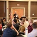 A speaker dressed in a black polo shirt stands at the front of a conference or seminar room, holding papers and addressing a seated audience. The room contains projection screens on both sides displaying presentation content, and an American flag is visible near the rear door. The audience appears to be a mixed group of young adults seated in rows of chairs.
