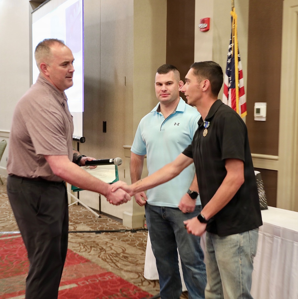 Three men are gathered in what appears to be a formal ceremony or recognition event held in a hotel ballroom or conference room. A man on the left is shaking hands with a younger man on the right who is wearing a medal, while a third man in a light blue polo shirt stands between them observing. An American flag is visible in the background alongside a projection screen.