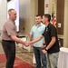 Three men are gathered in what appears to be a formal ceremony or recognition event held in a hotel ballroom or conference room. A man on the left is shaking hands with a younger man on the right who is wearing a medal, while a third man in a light blue polo shirt stands between them observing. An American flag is visible in the background alongside a projection screen.