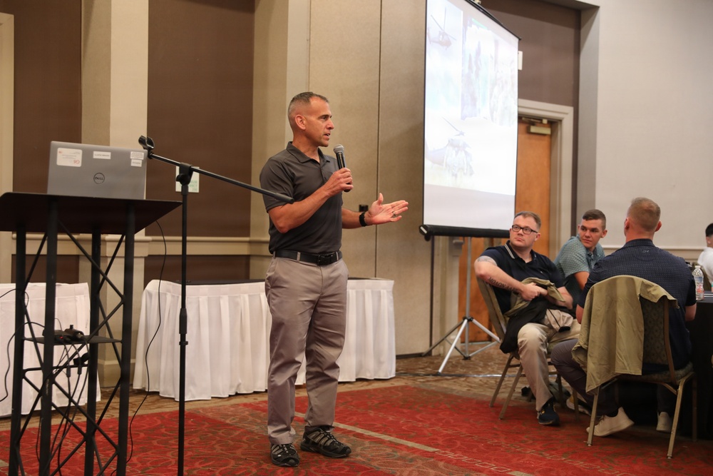 A man in a gray polo shirt is presenting at what appears to be a professional conference or seminar in a hotel ballroom setting. He is holding a microphone and gesturing with his hands while speaking to an audience. A projection screen displaying images is visible in the background, along with a Dell laptop on a podium.
