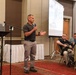 A man in a gray polo shirt is presenting at what appears to be a professional conference or seminar in a hotel ballroom setting. He is holding a microphone and gesturing with his hands while speaking to an audience. A projection screen displaying images is visible in the background, along with a Dell laptop on a podium.