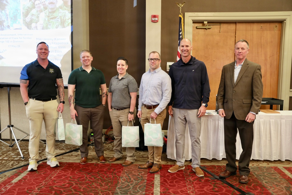 Six individuals are posed together in what appears to be a conference or banquet hall setting, with some holding gift bags. A presentation screen is visible in the background displaying text that includes names and what appears to be a military or organizational briefing. The group appears to be at a formal recognition or award ceremony event.