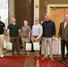 Six individuals are posed together in what appears to be a conference or banquet hall setting, with some holding gift bags. A presentation screen is visible in the background displaying text that includes names and what appears to be a military or organizational briefing. The group appears to be at a formal recognition or award ceremony event.