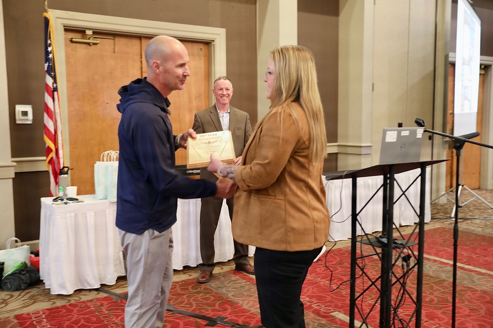 A formal award or recognition ceremony is taking place in what appears to be a hotel ballroom or conference room. A woman in a tan blazer is presenting a certificate or plaque to a bald man in a navy hoodie, while a third person in a sport coat smiles in the background. An American flag, a podium with a laptop, and draped tables are visible in the setting.