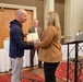 A formal award or recognition ceremony is taking place in what appears to be a hotel ballroom or conference room. A woman in a tan blazer is presenting a certificate or plaque to a bald man in a navy hoodie, while a third person in a sport coat smiles in the background. An American flag, a podium with a laptop, and draped tables are visible in the setting.