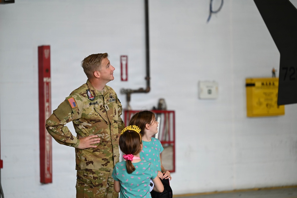 A U.S. military service member in OCP (Operational Camouflage Pattern) uniform stands in what appears to be an aircraft hangar alongside two young girls, one of whom is wearing a 'Happy Birthday' tiara. All three are looking upward, likely at an aircraft or display above them. The setting suggests a family day or open house event at a military installation.